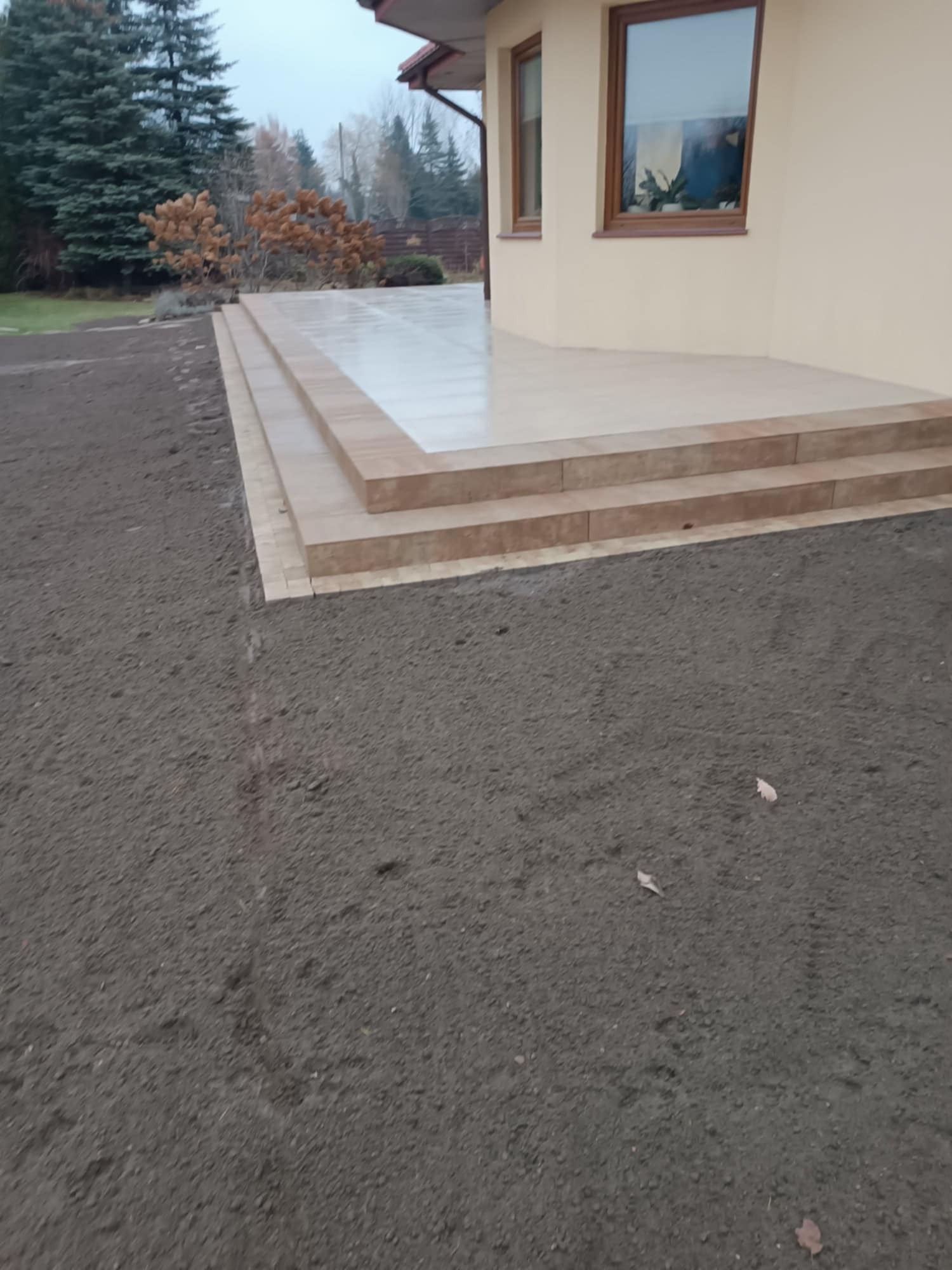 Wykończony taras z jasnych płytek, z trzema schodkami, przylegający do beżowej ściany domu. W tle widoczne drzewa i krzewy, a na pierwszym planie przygotowany teren pod trawnik.
