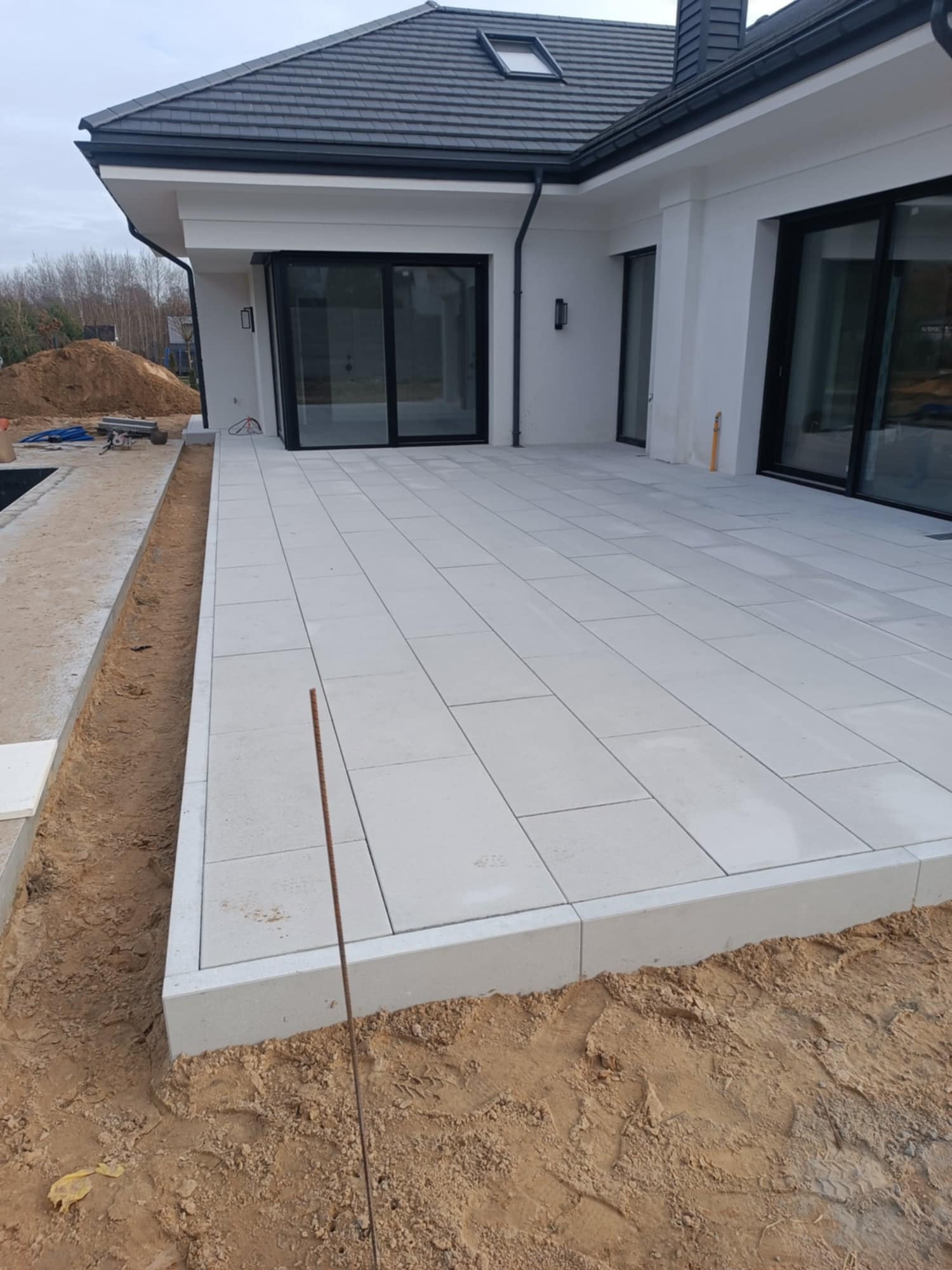 Nowoczesny taras z jasnoszarej, prostokątnej kostki brukowej, obrzeża z tego samego materiału. W tle dom z dużymi, przesuwnymi oknami i sterta ziemi.