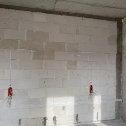Electro House Patryk Kobus - Surowa ściana z betonu komórkowego z poprowadzonymi przewodami elektrycznymi w białych listwach i puszkami instalacyjnymi z wystającymi kablami.