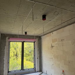 Electro House Patryk Kobus - Surowy pokój z widokiem na zieleń przez okno. Na suficie widoczne czarne oprawy oświetleniowe z czerwonymi detalami i poprowadzone kable elektryczne.