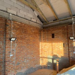 Electro House Patryk Kobus - Surowa ściana z cegły z zamontowanym oświetleniem zewnętrznym i przewodami w białych listwach. Widoczne drewniane elementy konstrukcji dachu.