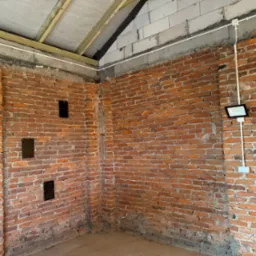 Surowe wnętrze z ceglanymi ścianami, gdzie zamontowano dwa reflektory LED z białymi kablami w peszlu. Widoczne otwory na instalacje i drewniana konstrukcja dachu.