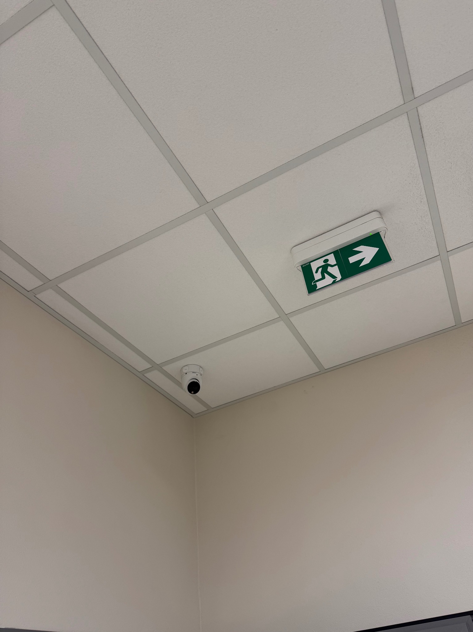 Biały sufit kasetonowy z zamontowaną kamerą monitoringu i podświetlanym znakiem ewakuacyjnym ze strzałką w prawo i piktogramem biegnącej osoby.