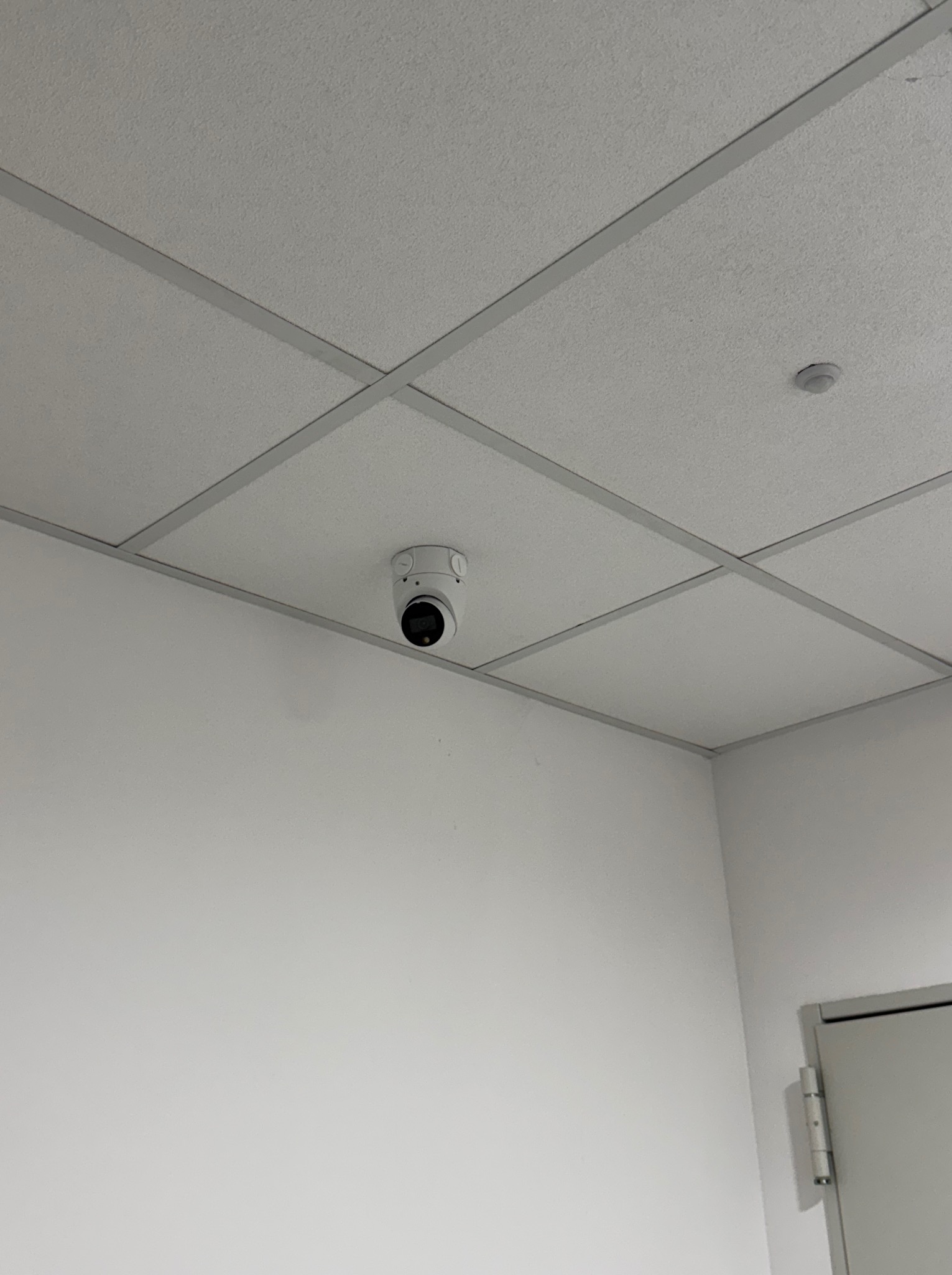 Biała kamera monitoringu kopułkowa zamontowana na suficie kasetonowym w jasnym pomieszczeniu, widoczny fragment drzwi po prawej.