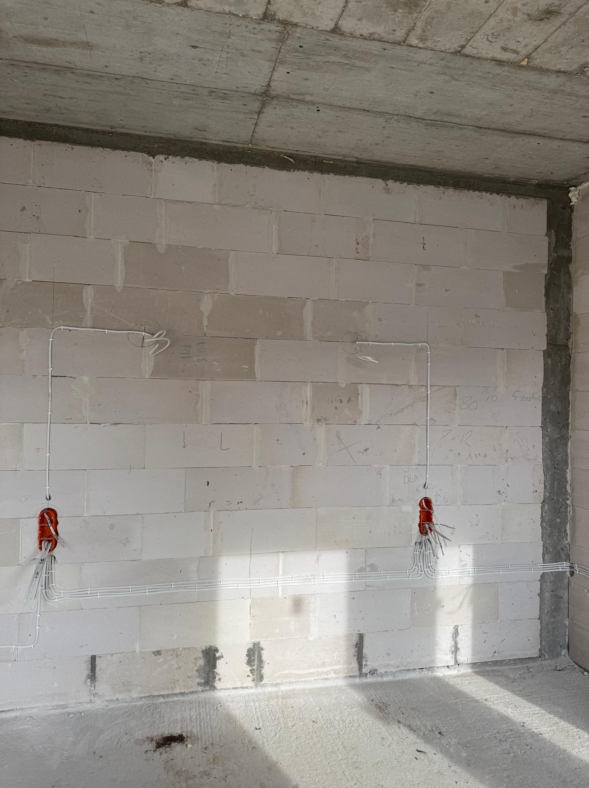 Surowa ściana z betonu komórkowego z poprowadzonymi przewodami elektrycznymi w białych listwach i puszkami instalacyjnymi z wystającymi kablami.