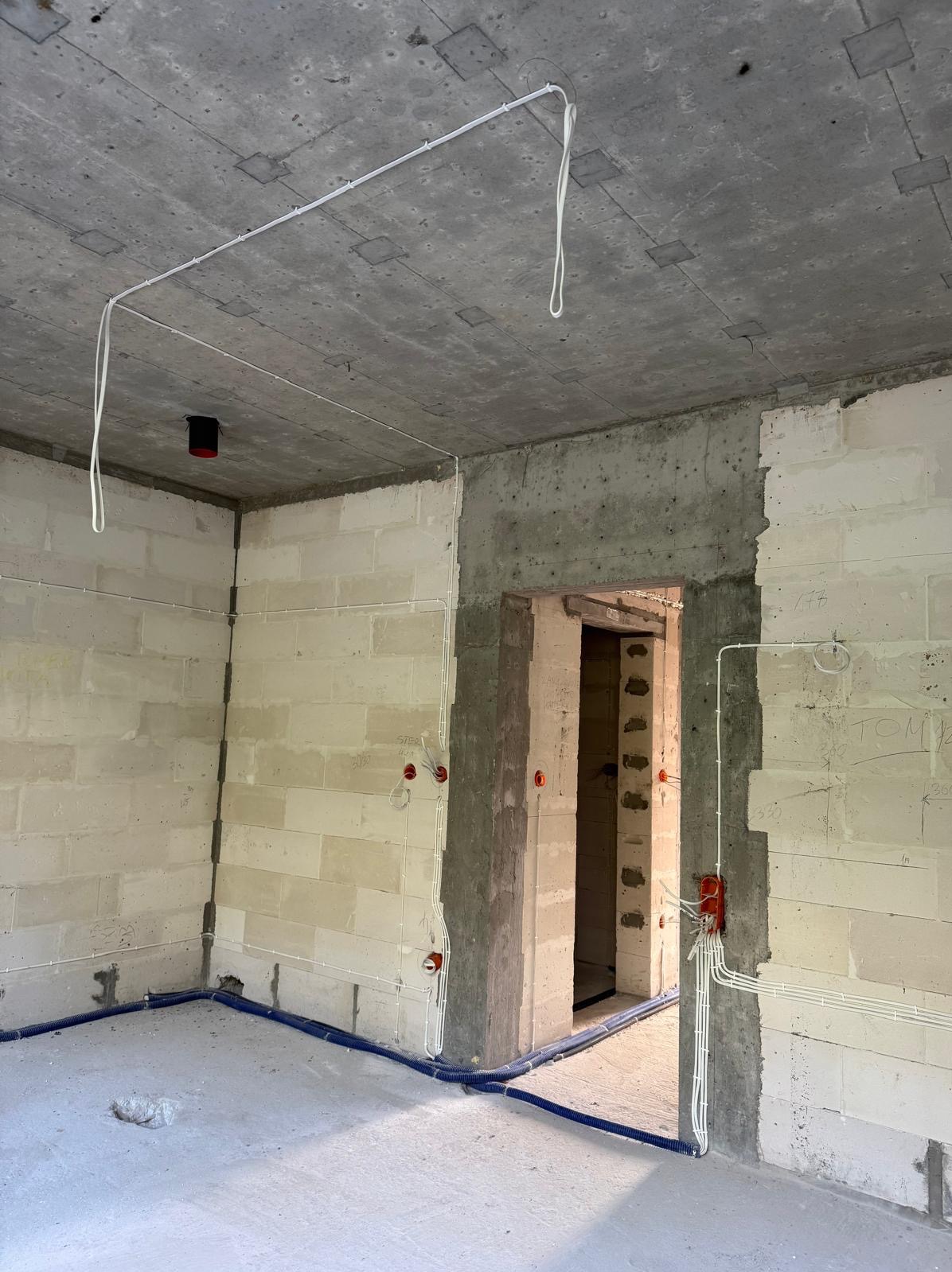 Surowy stan: Instalacja elektryczna w budynku z pustaków, z widocznymi przewodami podtynkowymi i rurami ochronnymi na podłodze. Otwory pod gniazdka i włączniki.