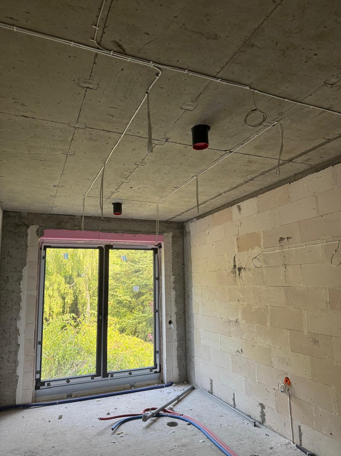 Surowy pokój z widokiem na zieleń przez okno. Na suficie widoczne czarne oprawy oświetleniowe z czerwonymi detalami i poprowadzone kable elektryczne.