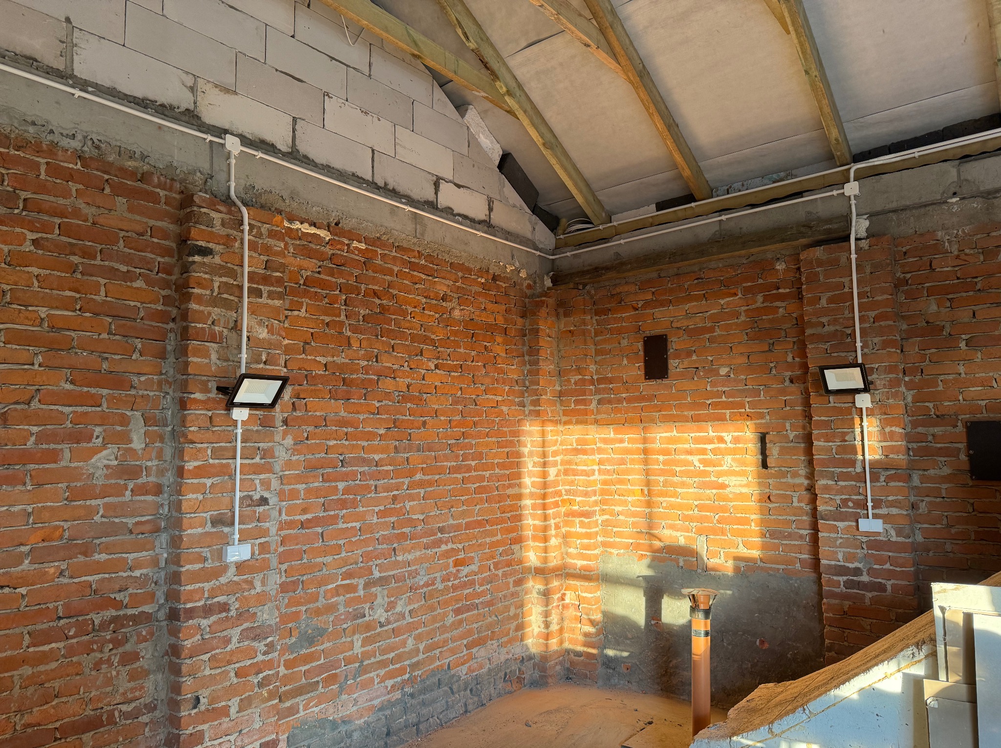 Surowa ściana z cegły z zamontowanym oświetleniem zewnętrznym i przewodami w białych listwach. Widoczne drewniane elementy konstrukcji dachu.