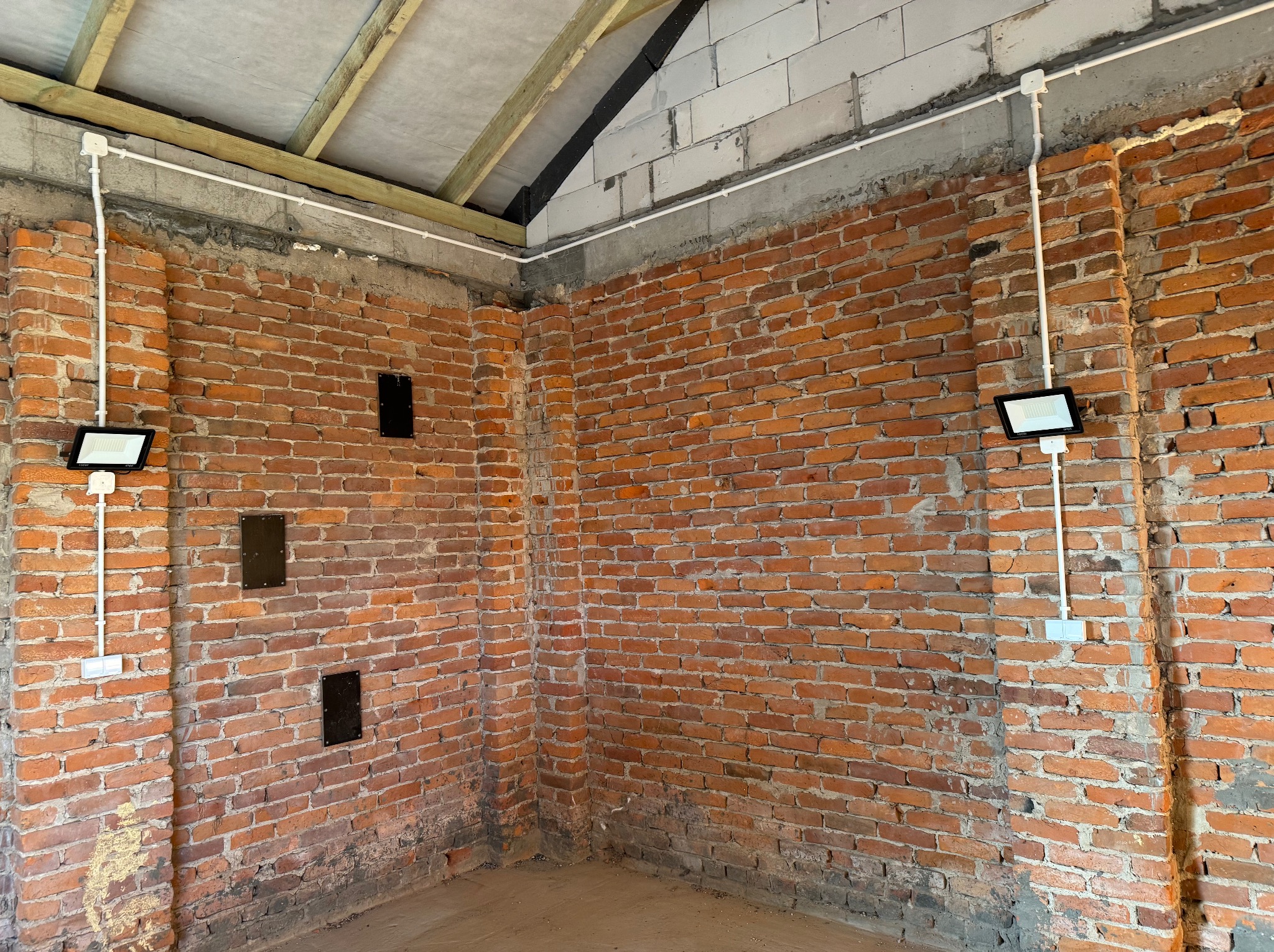 Surowe wnętrze z ceglanymi ścianami, gdzie zamontowano dwa reflektory LED z białymi kablami w peszlu. Widoczne otwory na instalacje i drewniana konstrukcja dachu.