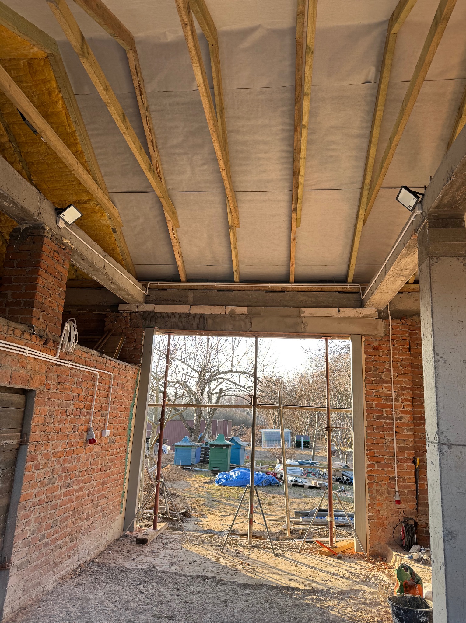 Wnętrze w surowym stanie z widoczną instalacją elektryczną, oświetleniem i izolacją poddasza. Ceglana ściana z przewodami i gniazdem. W tle widok na zewnątrz.