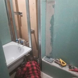 Budowa OSG Paweł Rutkowski - Montaż instalacji wodno-kanalizacyjnej w łazience. Widoczna wanna, metalowa konstrukcja pod płyty gipsowe, narzędzia oraz osoba w czerwonej koszuli flanelowej przy pracy.