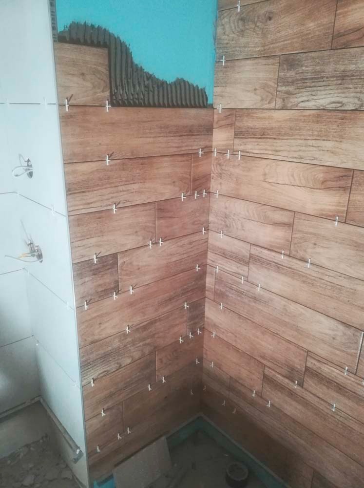 Układanie płytek imitujących drewno w rogu łazienki, widoczne krzyżyki dystansowe i nałożony klej na ścianie. Częściowo wykończona ściana z białych płytek.