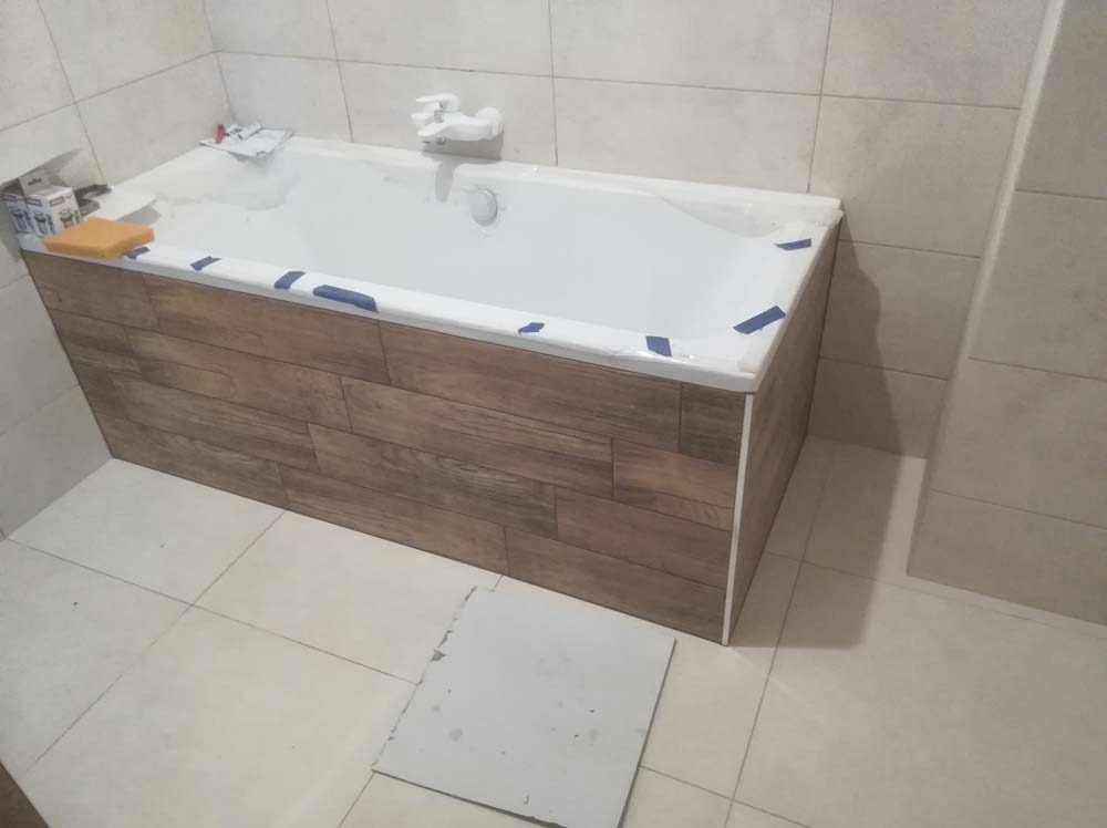 Obudowa wanny wykończona panelami imitującymi drewno w łazience z białymi płytkami. Widoczne niebieskie taśmy zabezpieczające podczas montażu i akcesoria łazienkowe.