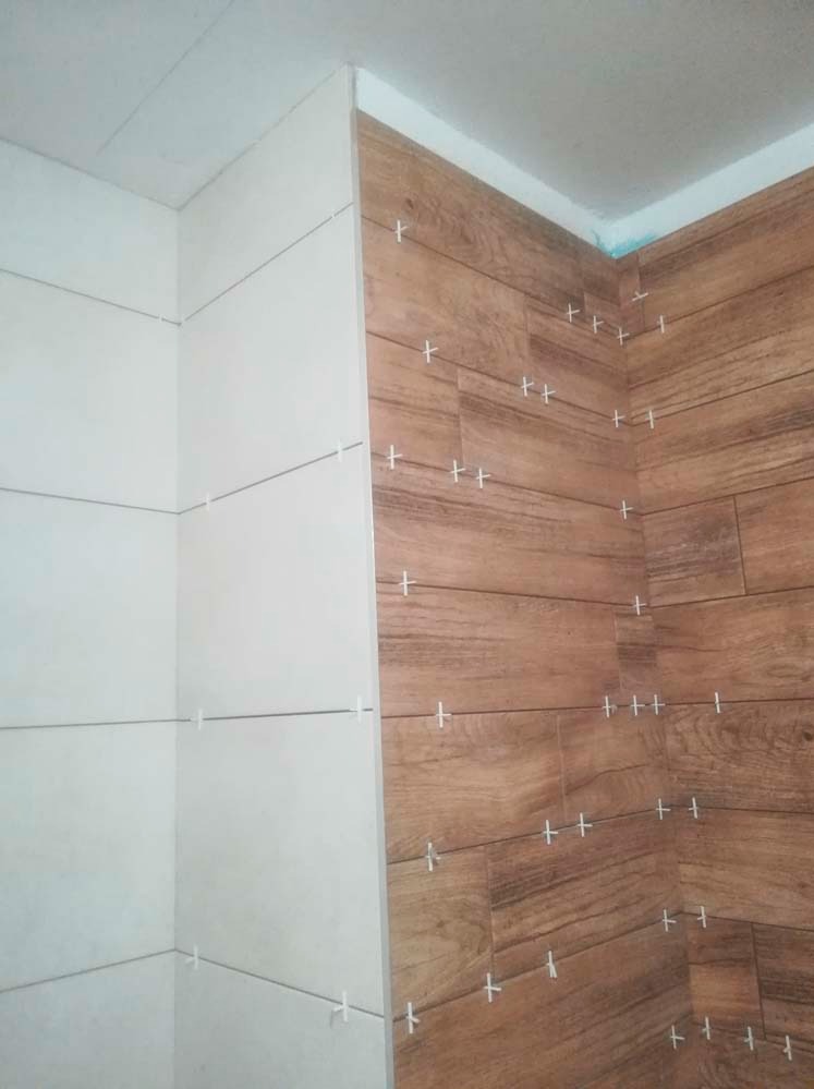Ściana wyłożona dwoma rodzajami płytek: białe, poziome płytki i brązowe płytki imitujące drewno. Widoczne krzyżyki dystansowe. Ukończony etap prac w łazience.