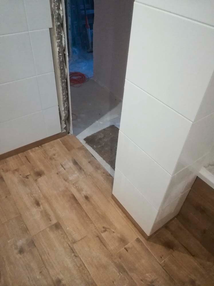 Wykończona łazienka z drewnopodobną podłogą i białymi, kwadratowymi płytkami na ścianach. Widoczny fragment niedokończonego przejścia do innego pomieszczenia.