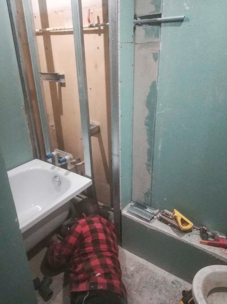 Montaż instalacji wodno-kanalizacyjnej w łazience. Widoczna wanna, metalowa konstrukcja pod płyty gipsowe, narzędzia oraz osoba w czerwonej koszuli flanelowej przy pracy.