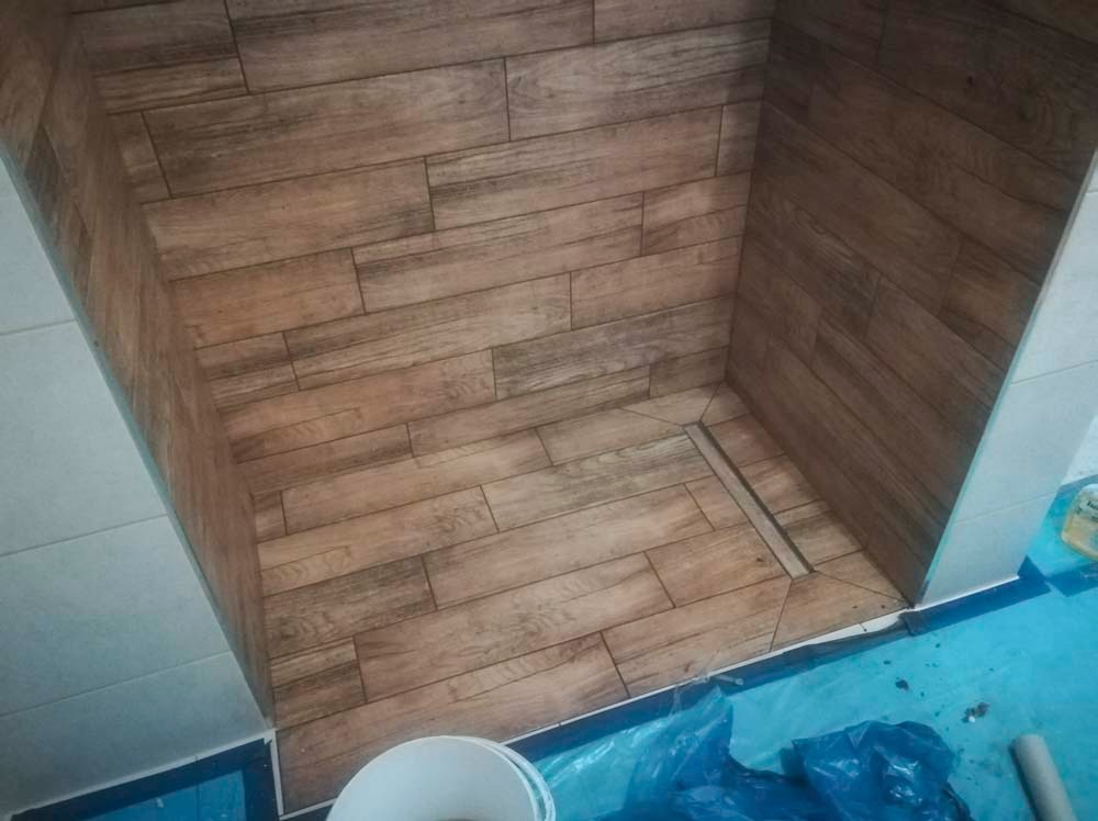 Wykończona kabina prysznicowa z drewnopodobnymi płytkami ułożonymi horyzontalnie na ścianach i podłodze, z odpływem liniowym. Widoczne białe płytki na zewnątrz kabiny.