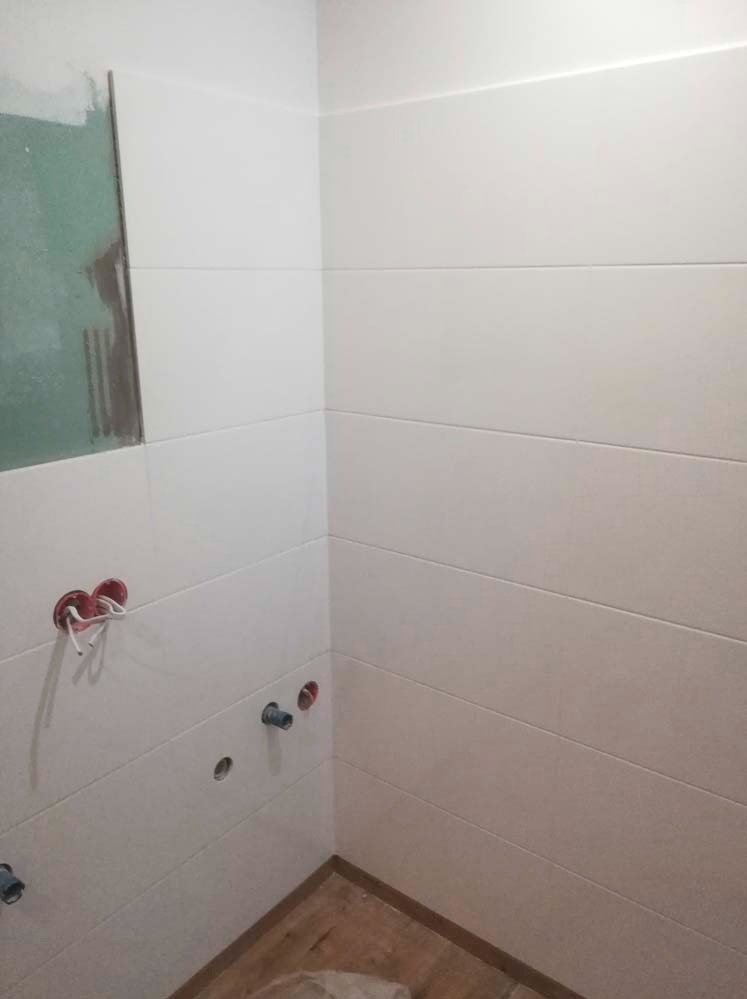 Wykończona łazienka z poziomymi, białymi płytkami. Widoczne przygotowane instalacje wodne i elektryczne. Fragment ściany z zieloną płytą kartonowo-gipsową.