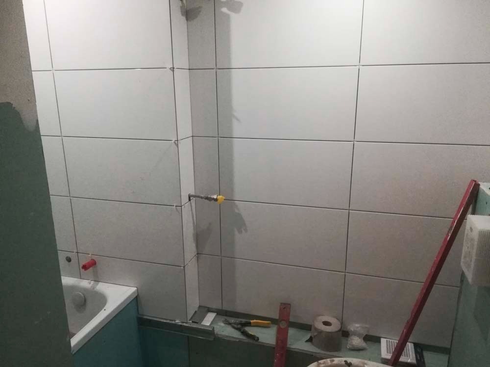 Białe płytki ścienne w łazience, częściowo ułożone, widoczne narzędzia glazurnicze i fragment wanny. Widok na ścianę z instalacją wodną.