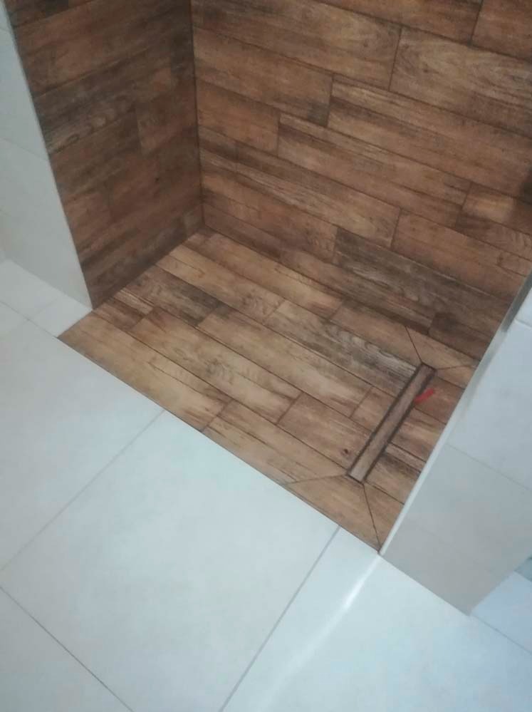 Wykończony brodzik z odpływem liniowym, wyłożony płytkami imitującymi drewno, połączony z białą podłogą. Estetyczne i funkcjonalne rozwiązanie.