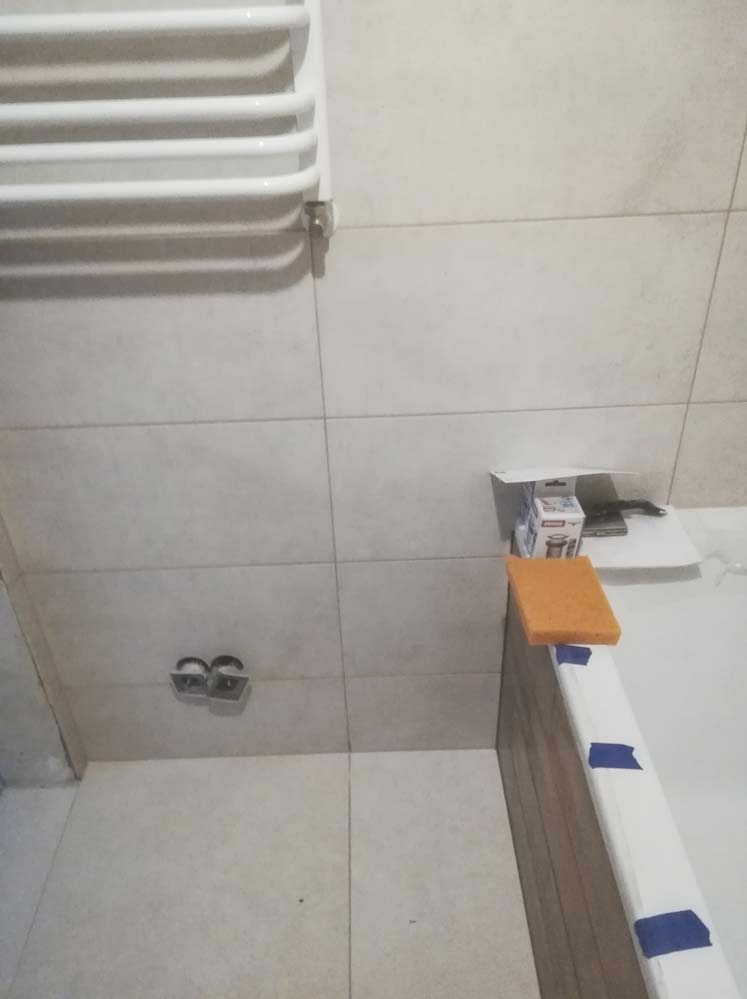Białe płytki w łazience z grzejnikiem drabinkowym, przygotowane do fugowania. Na wannie leży gąbka, widoczne akcesoria i otwory na armaturę.