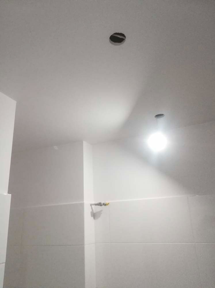 Białe wnętrze z otworem w suficie i widoczną instalacją. Sufit pomalowany na biało, płytki na ścianach. Widoczny fragment instalacji. Ujęcie z dołu.