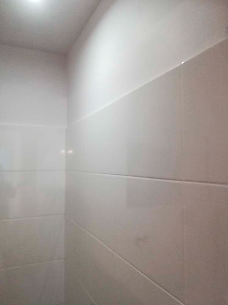 Ściana wyłożona błyszczącymi, białymi płytkami ceramicznymi w łazience. Widoczny narożnik pomieszczenia i oświetlenie sufitowe.