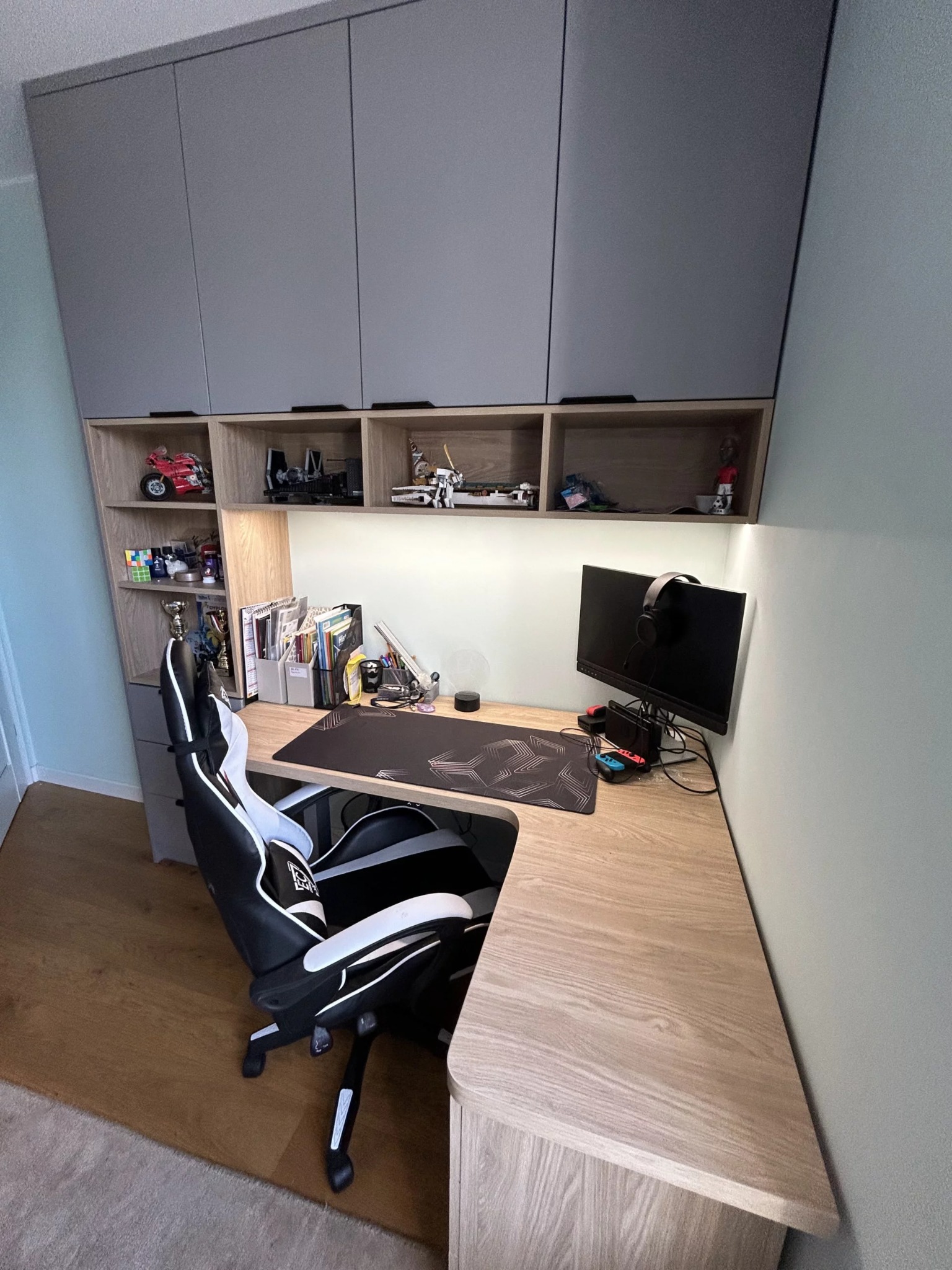 Ergonomiczne biurko narożne z szarymi szafkami wiszącymi, oświetleniem LED i półkami. Krzesło gamingowe, monitor i akcesoria tworzą funkcjonalną przestrzeń.