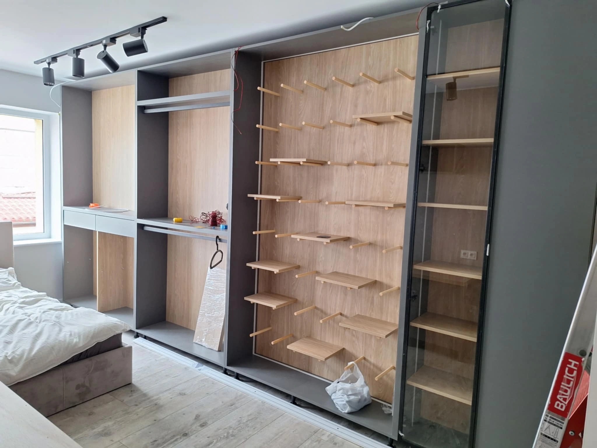 Szafa wnękowa z drewnianymi półkami i szklanymi drzwiami w Gdańsku. Widoczny fragment łóżka i oświetlenie sufitowe. Montaż w trakcie realizacji.