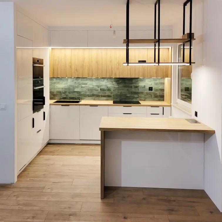Nowoczesna kuchnia z białymi szafkami, drewnianymi blatami i zieloną mozaiką. Nad wyspą kuchenną designerska lampa z drewnianymi elementami.