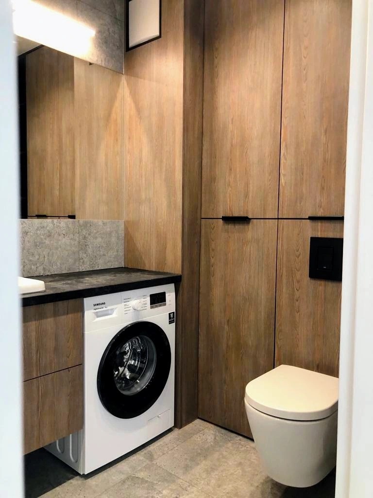Funkcjonalna łazienka z zabudową meblową w odcieniach drewna, zintegrowaną pralką Samsung i podwieszaną toaletą. Minimalistyczny design, szare płytki, oświetlenie LED.