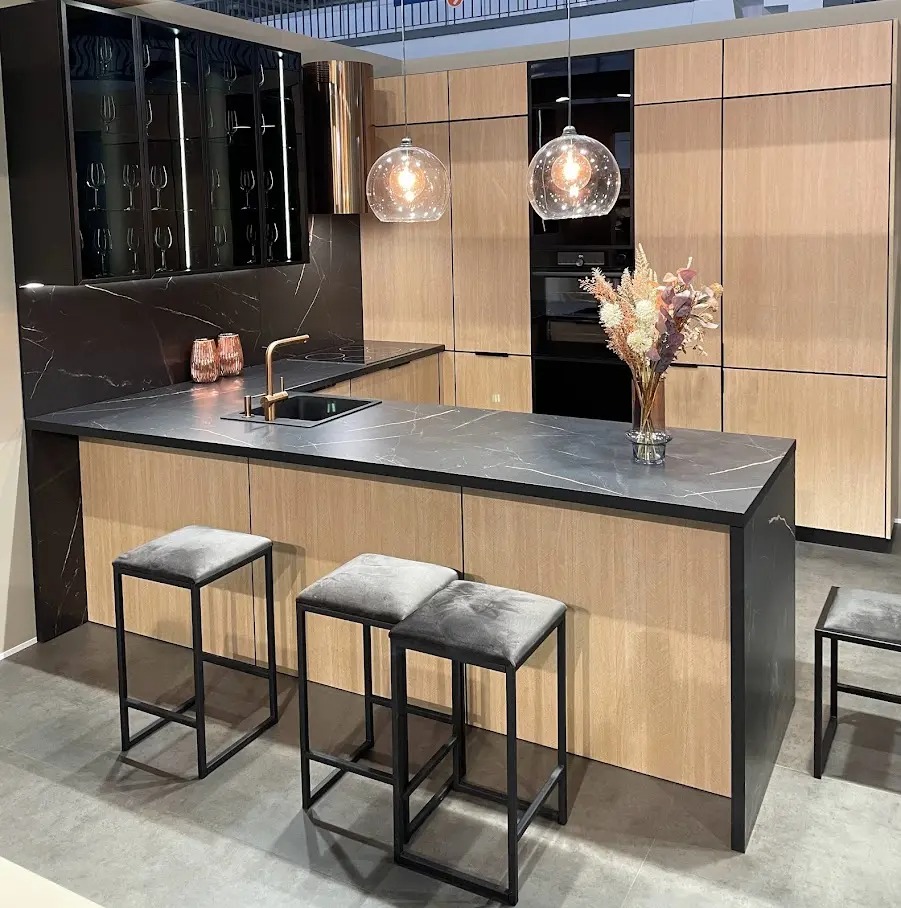 Nowoczesna kuchnia z wyspą, blatem z czarnego marmuru i drewnianymi frontami. Nad wyspą wiszą dwie szklane lampy. Ustawione są trzy szare hokery.