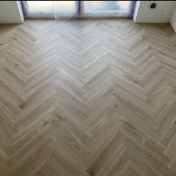 Dominik Dzięgiel - Ułożona podłoga z paneli w jodełkę, jasne drewno, widok z góry, okno zabezpieczone folią malarską, ściany przygotowane do malowania, gniazdko elektryczne.