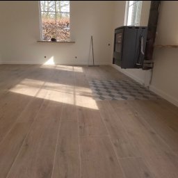 Dominik Dzięgiel - Jasne, drewnopodobne płytki podłogowe w nowym wnętrzu z piecem wolnostojącym. Fragment dekoracyjnej mozaiki. Okno z widokiem na drzewa. Słoneczne światło.