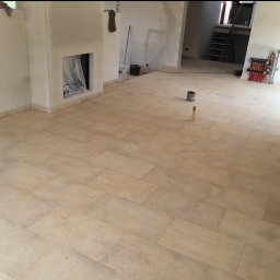 Dominik Dzięgiel - Jasne, beżowe płytki podłogowe ułożone w przestronnym wnętrzu z przygotowanym kominkiem i instalacją grzewczą. Widoczne ślady prac wykończeniowych.