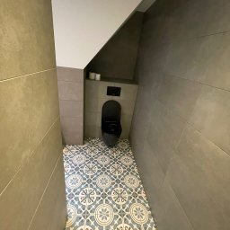 Dominik Dzięgiel - Wąska łazienka z szarą ceramiką ścienną i wzorzystą, błękitno-białą podłogą. Podwieszana toaleta w zabudowie. Widok z góry podkreśla unikalny układ pomieszczenia.