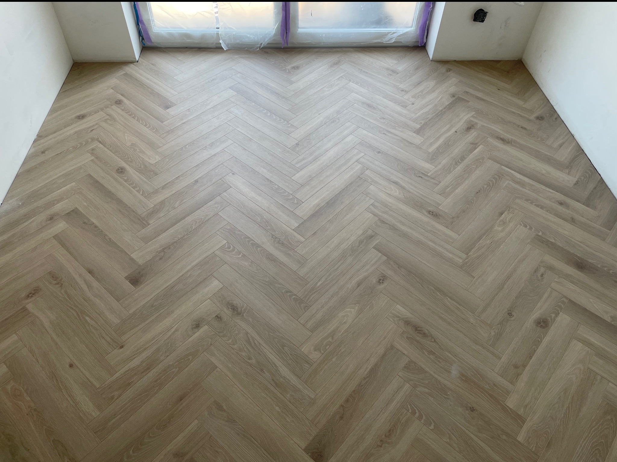 Ułożona podłoga z paneli w jodełkę, jasne drewno, widok z góry, okno zabezpieczone folią malarską, ściany przygotowane do malowania, gniazdko elektryczne.