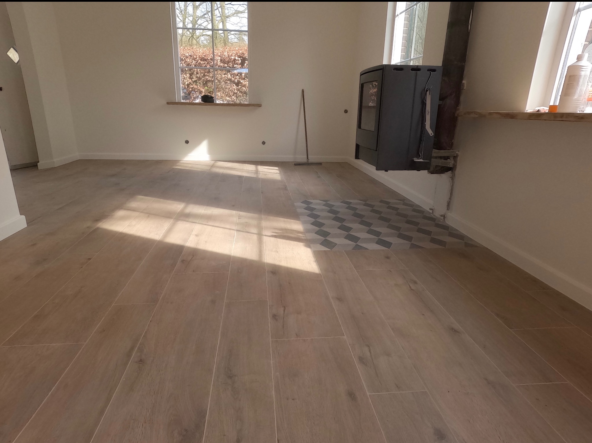 Jasne, drewnopodobne płytki podłogowe w nowym wnętrzu z piecem wolnostojącym. Fragment dekoracyjnej mozaiki. Okno z widokiem na drzewa. Słoneczne światło.