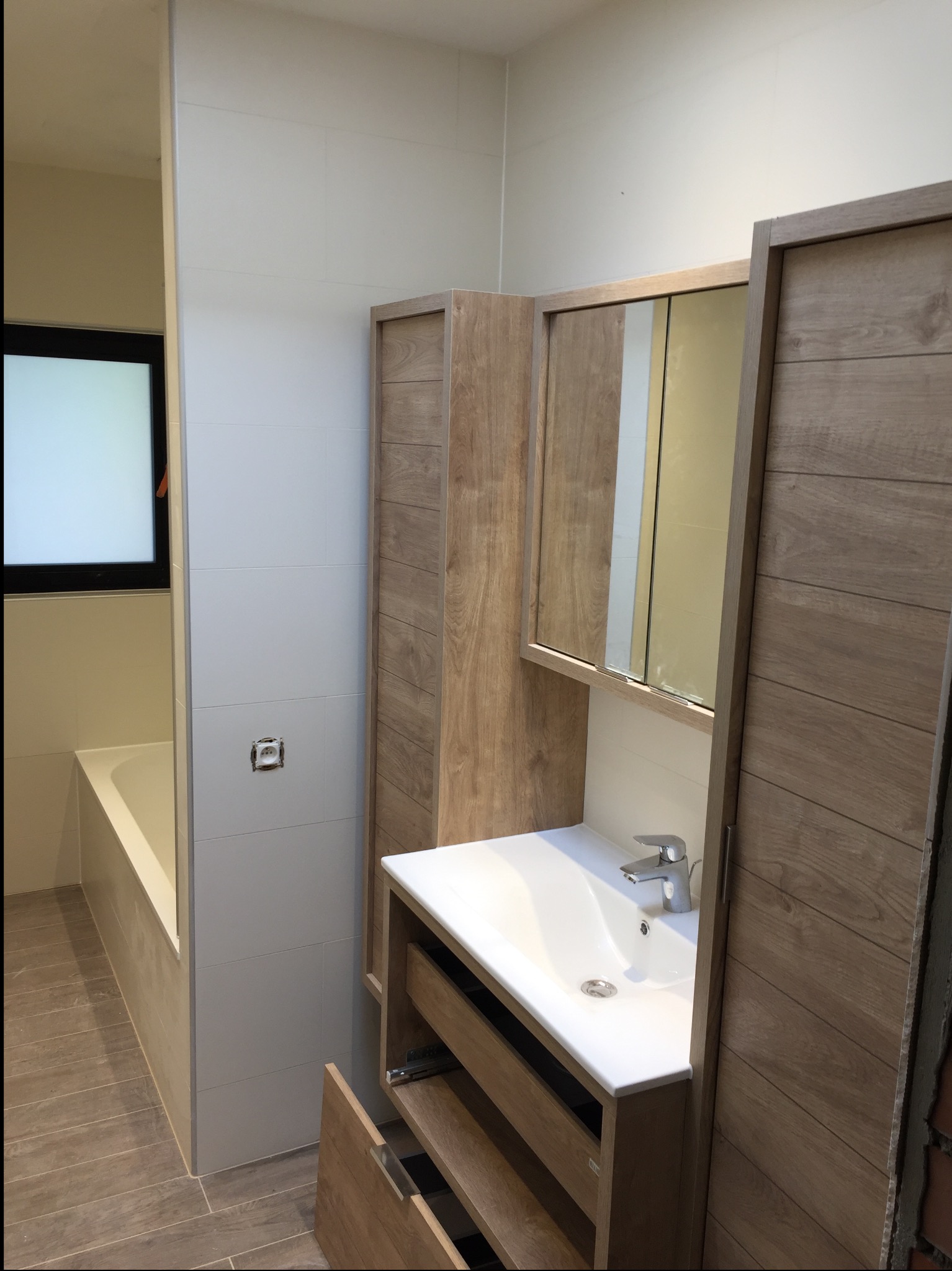 Jasna łazienka z drewnianymi szafkami, białą umywalką i wanną. Widoczne płytki na ścianach i podłodze. Otwarta szuflada szafki pod umywalką.