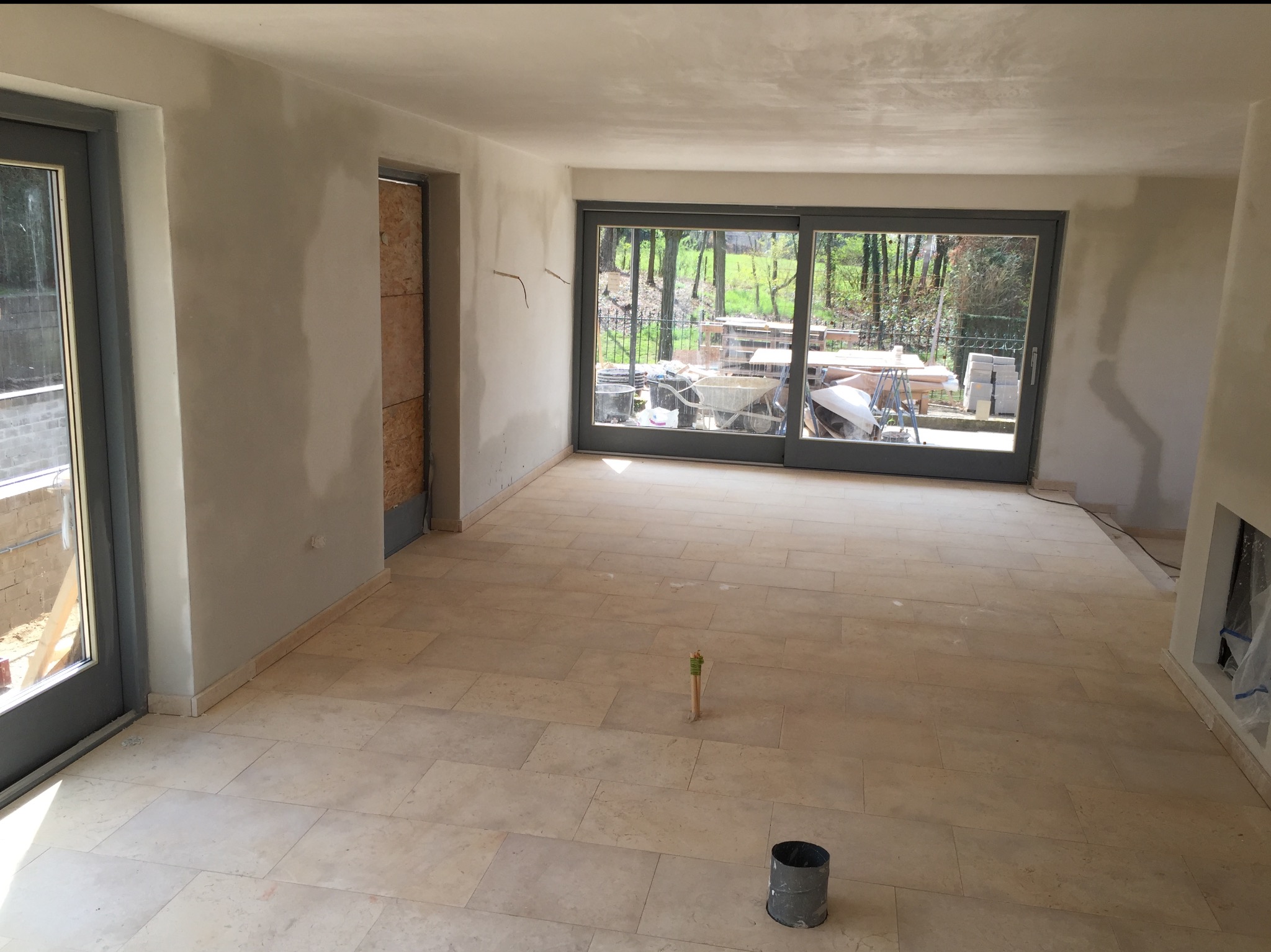 Wnętrze w trakcie prac: świeżo położona jasna podłoga z płytek, ściany po szpachlowaniu, duże okno z widokiem na zieleń. Widoczne narzędzia i materiały budowlane.