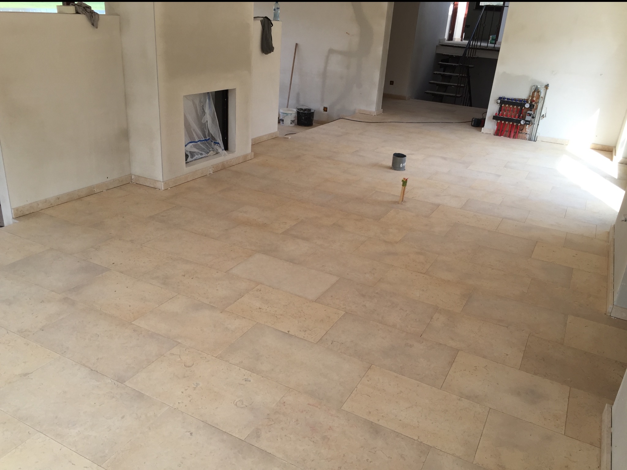 Jasne, beżowe płytki podłogowe ułożone w przestronnym wnętrzu z przygotowanym kominkiem i instalacją grzewczą. Widoczne ślady prac wykończeniowych.