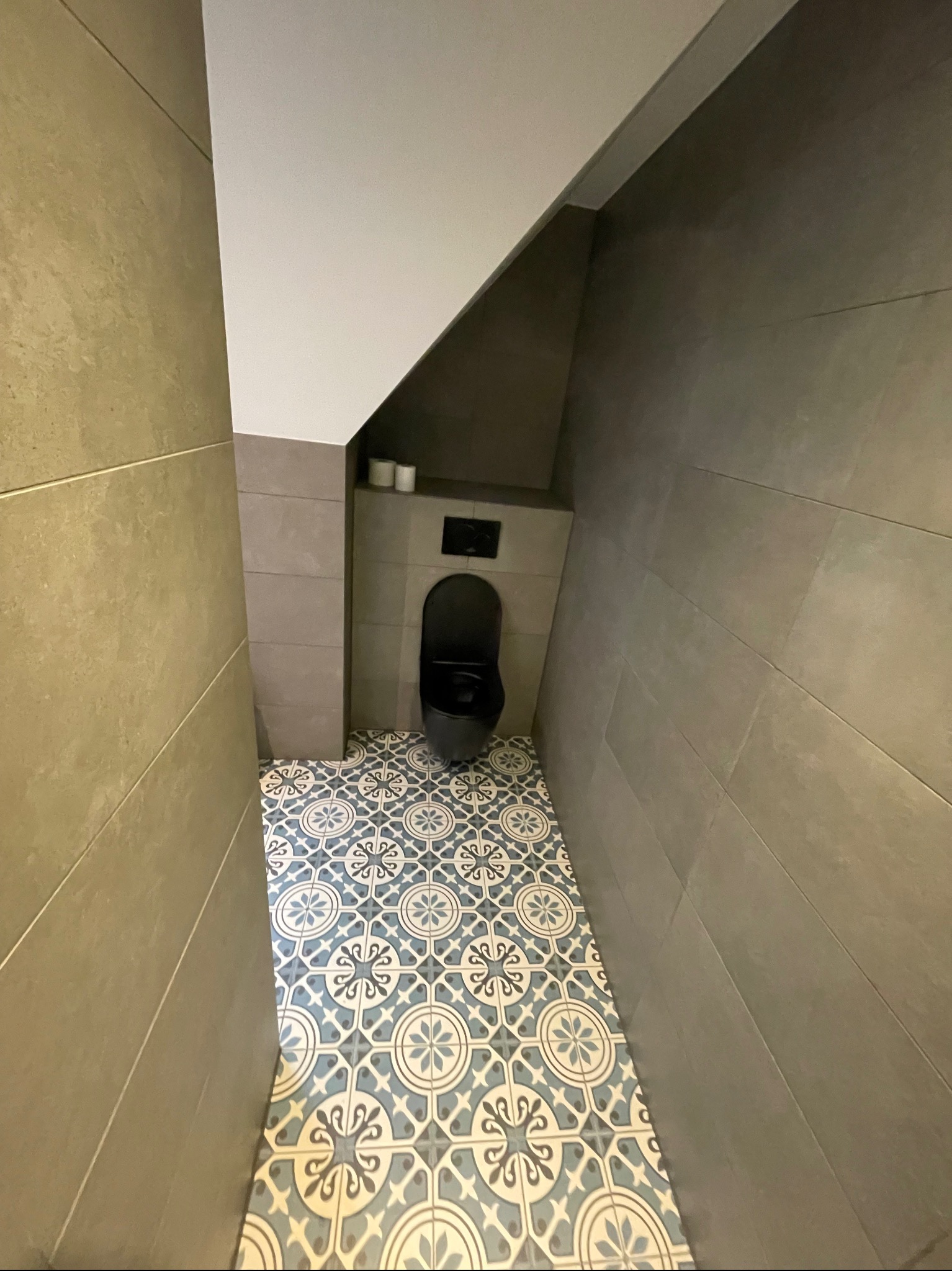 Wąska łazienka z szarą ceramiką ścienną i wzorzystą, błękitno-białą podłogą. Podwieszana toaleta w zabudowie. Widok z góry podkreśla unikalny układ pomieszczenia.