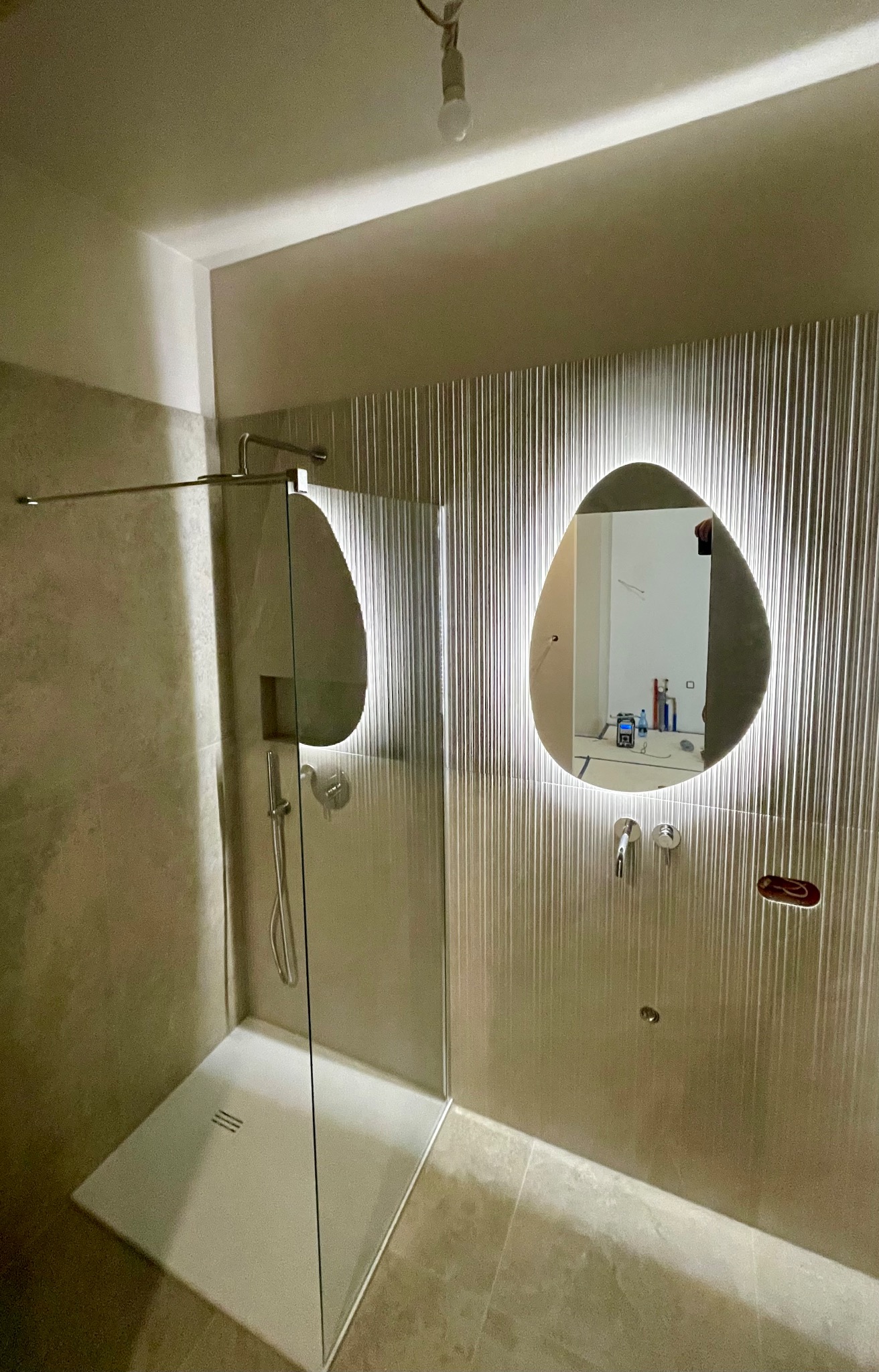 Nowoczesna kabina prysznicowa z owalnym lustrem podświetlanym LED, wykończona płytkami w odcieniach beżu z delikatnym wzorem. Minimalistyczny design, widoczne baterie.