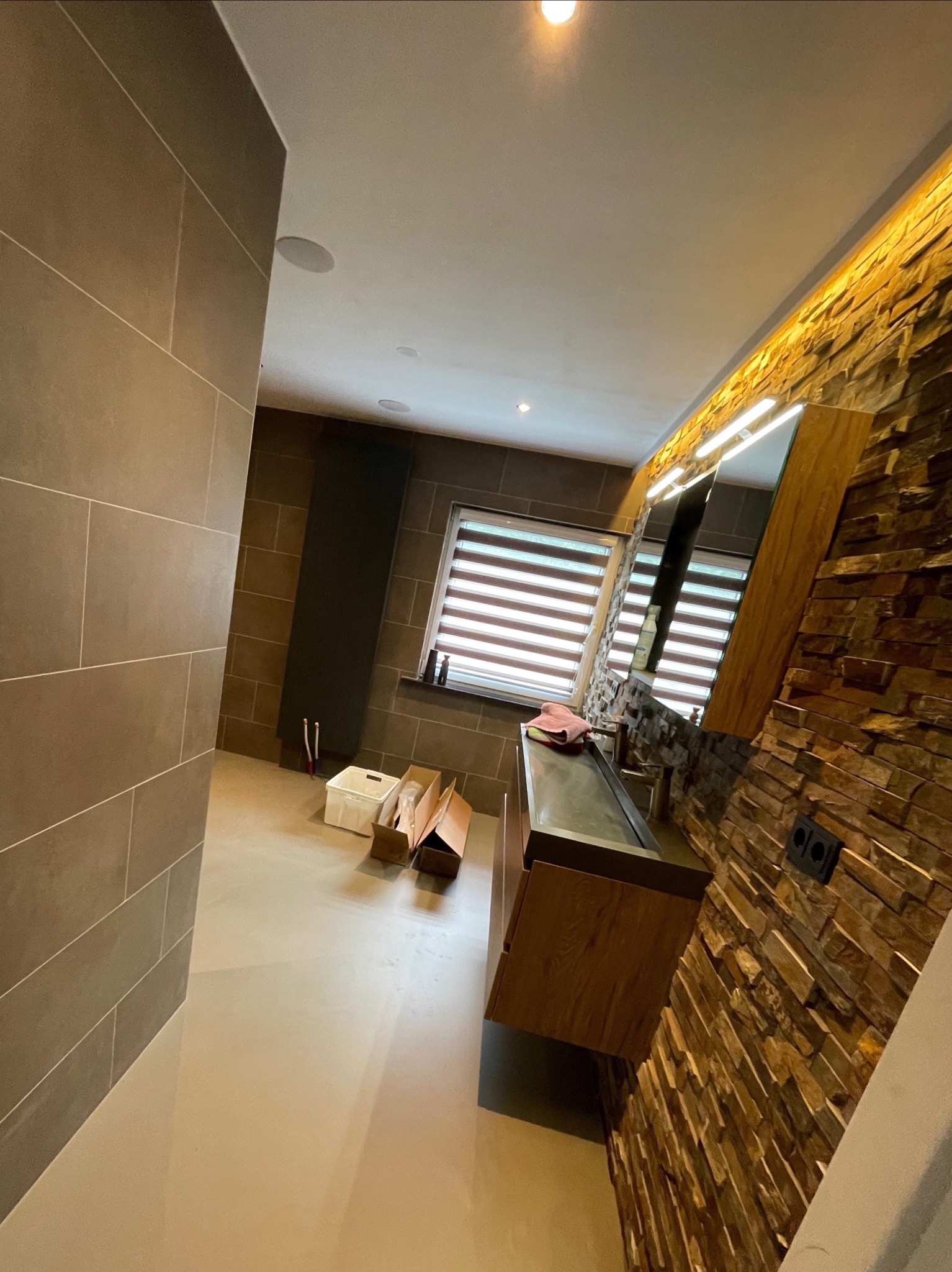 Nowoczesna łazienka z kamienną ścianą, drewnianą szafką pod umywalką i dużym lustrem z oświetleniem LED. Widoczne płytki w odcieniach brązu i beżu.