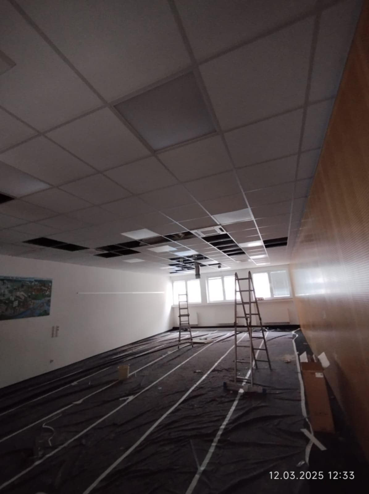 Montaż sufitu podwieszanego rastrowego. Widoczne drabiny, zabezpieczona podłoga folią i częściowo zamontowane płyty sufitowe. Oświetlenie sufitowe w tle.