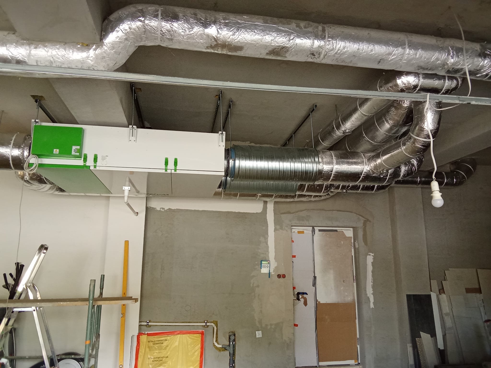 Szczegółowe ujęcie instalacji wentylacyjnej z aluminiowymi rurami pod sufitem w surowym wnętrzu. Widoczny rekuperator i elementy montażowe.