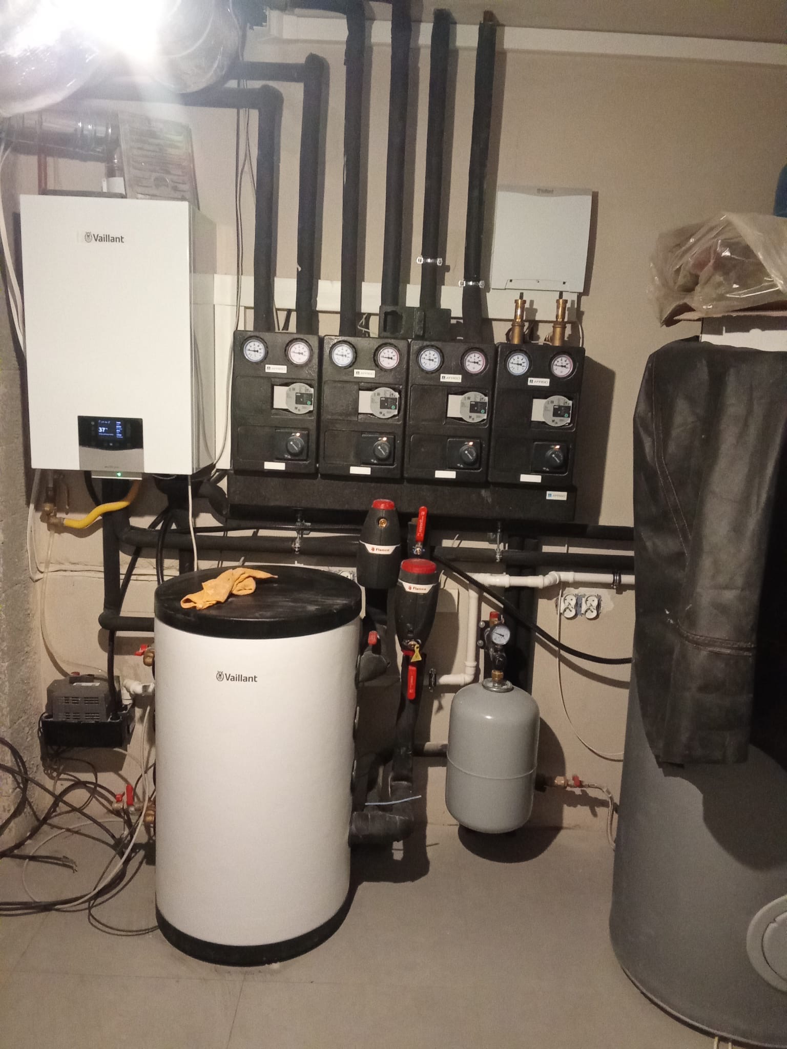 Kotłownia z kotłem gazowym Vaillant, zbiornikiem i rozdzielaczem. Widoczne czarne rury, manometry i zawory. Na zbiorniku leży pomarańczowa ścierka.