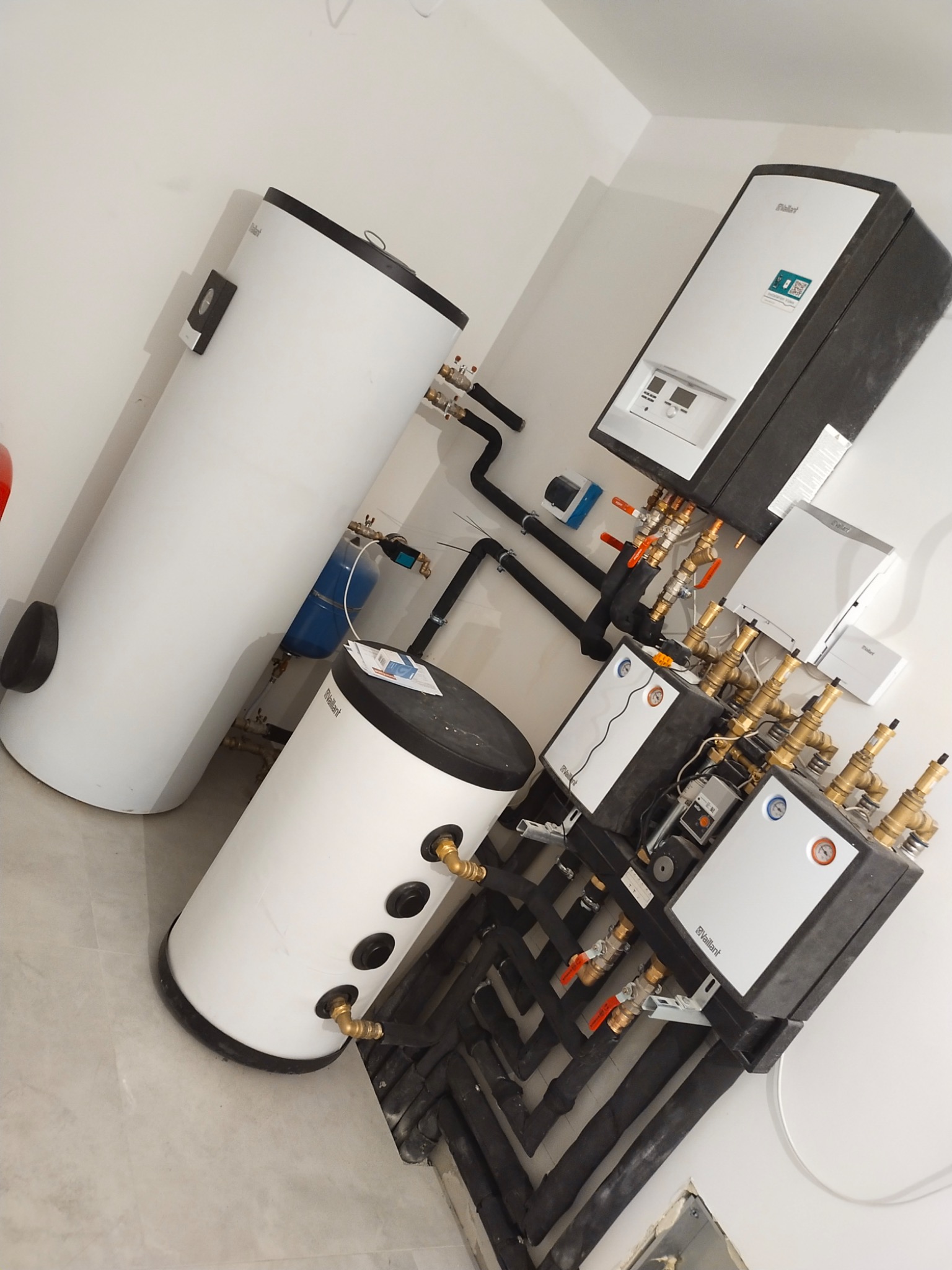 Kotłownia z pompą ciepła Vaillant: dwa białe zbiorniki, czarne rury i moduły sterujące z mosiężnymi zaworami, instalacja w domu jednorodzinnym, ujęcie pod kątem.