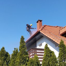 Krzysztof Kuliński - Pracownik w kasku i uprzęży czyści dach z czerwonej dachówki za pomocą myjki ciśnieniowej. W tle błękitne niebo i zielone tuje.