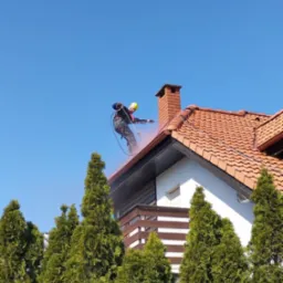 Pracownik w kasku i uprzęży czyści dach z czerwonej dachówki za pomocą myjki ciśnieniowej. W tle błękitne niebo i zielone tuje.